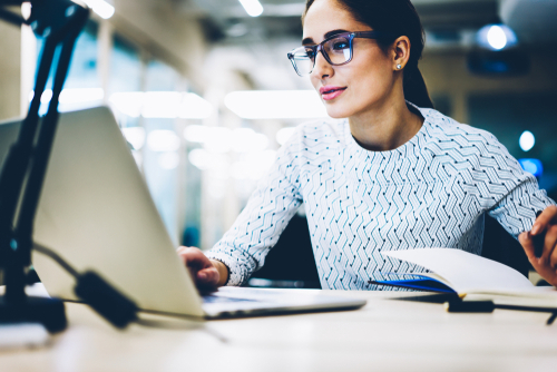 A woman in a light blue patterned shirt and glasses works on her laptop and takes notes in a notepad. 