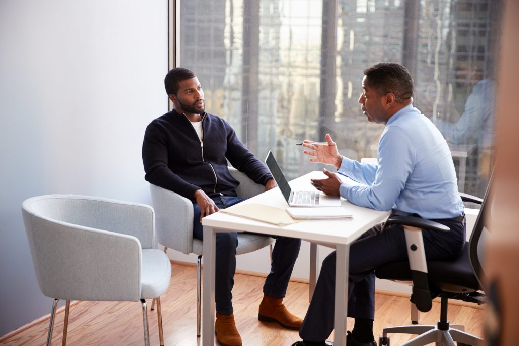 An African American business professional sits at his desk across from an African American client wearing a black sweater. 