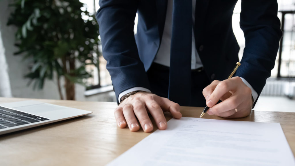 Small business owner in suit and tie leans on desk to sign Business Owner Policy (BOP) document, laptop computer nearby.
