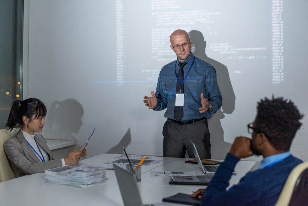 An IT professional delivers a presentation on cyber security best practices to a corporate team seated around a boardroom table.