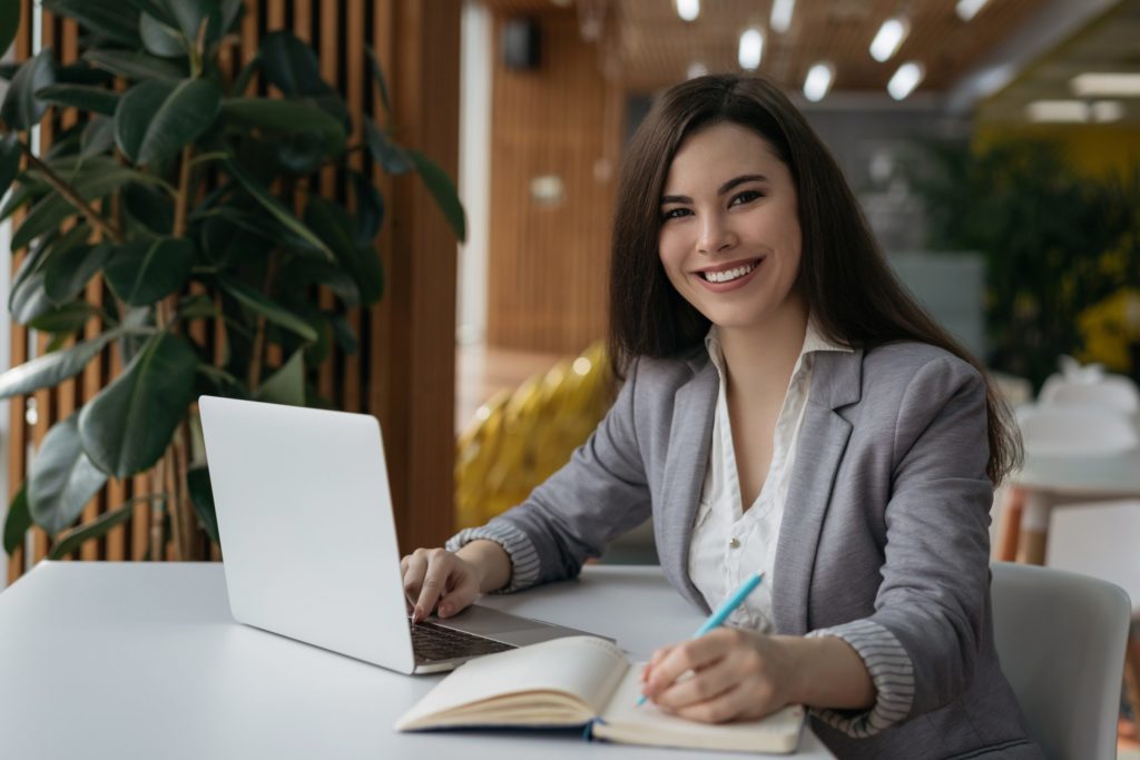Smiling insurance broker sits at table, researching cyber insurance policy options on laptop computer and writing in notebook.