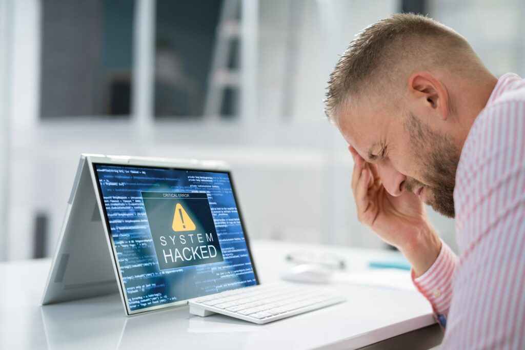 Businessman sits at desk, eyes closed, rubbing his forehead, as his folding laptop screen displays a “System Hacked” alert. 