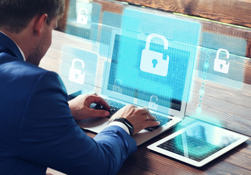 A young man in a suit sits at a table and types on his laptop while a blue graphic of a lock symbolizes cyber security. 