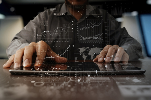 Close-up of a man in a grey shirt showing his hands as he works on a tablet with graphs & data graphics overlaying the image. 