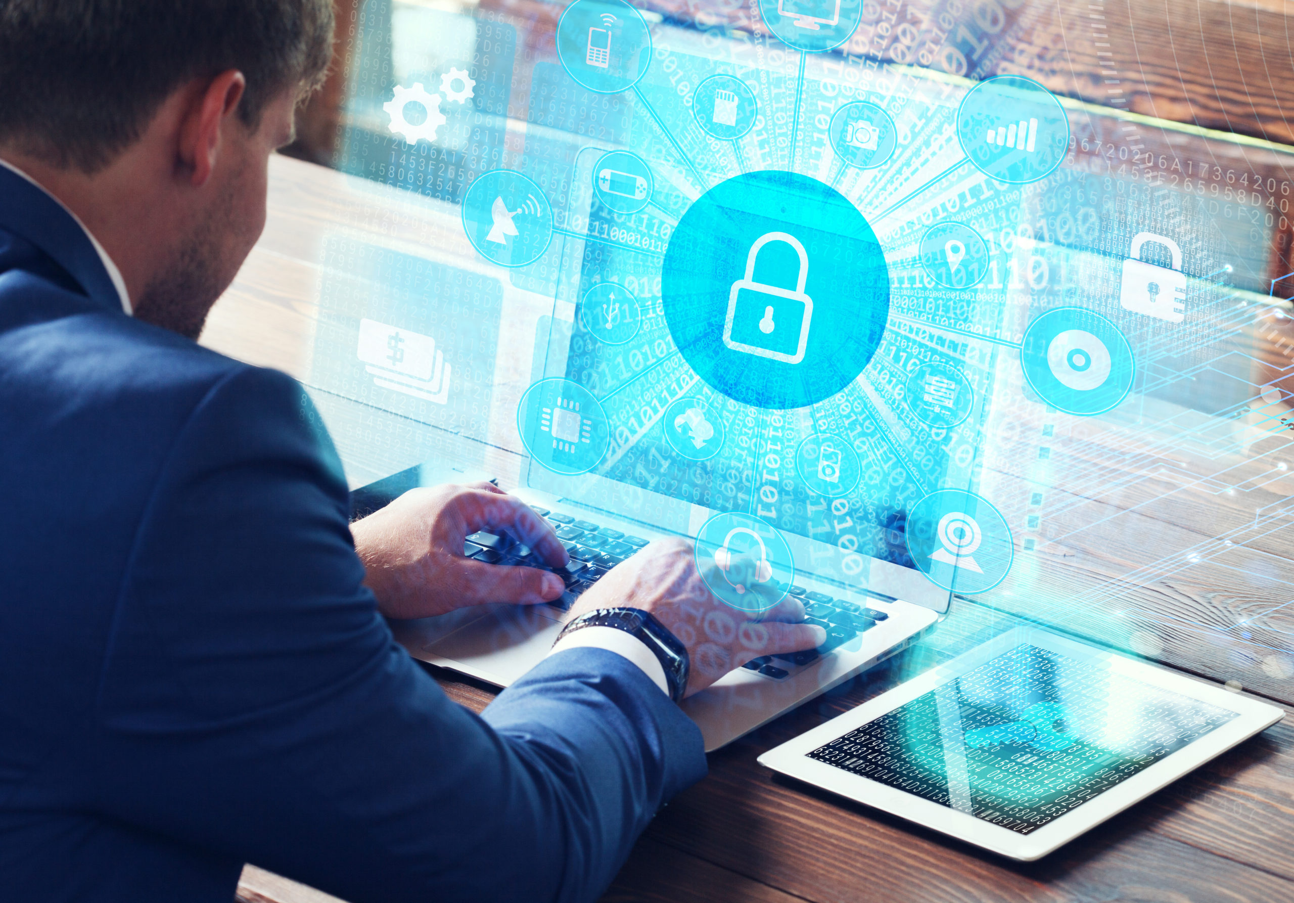 Businessman types on laptop computer, tablet computer nearby, overlaid with padlock icon illustrating cyber risk management.