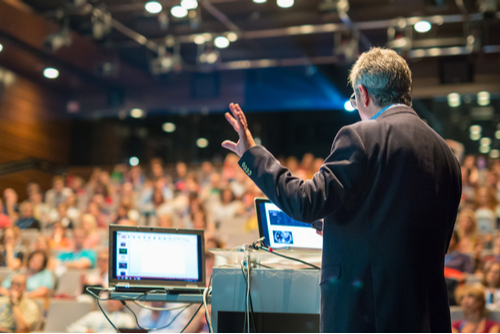 A man in a suit refers to his computer as he gives a presentation in front of a large audience. 